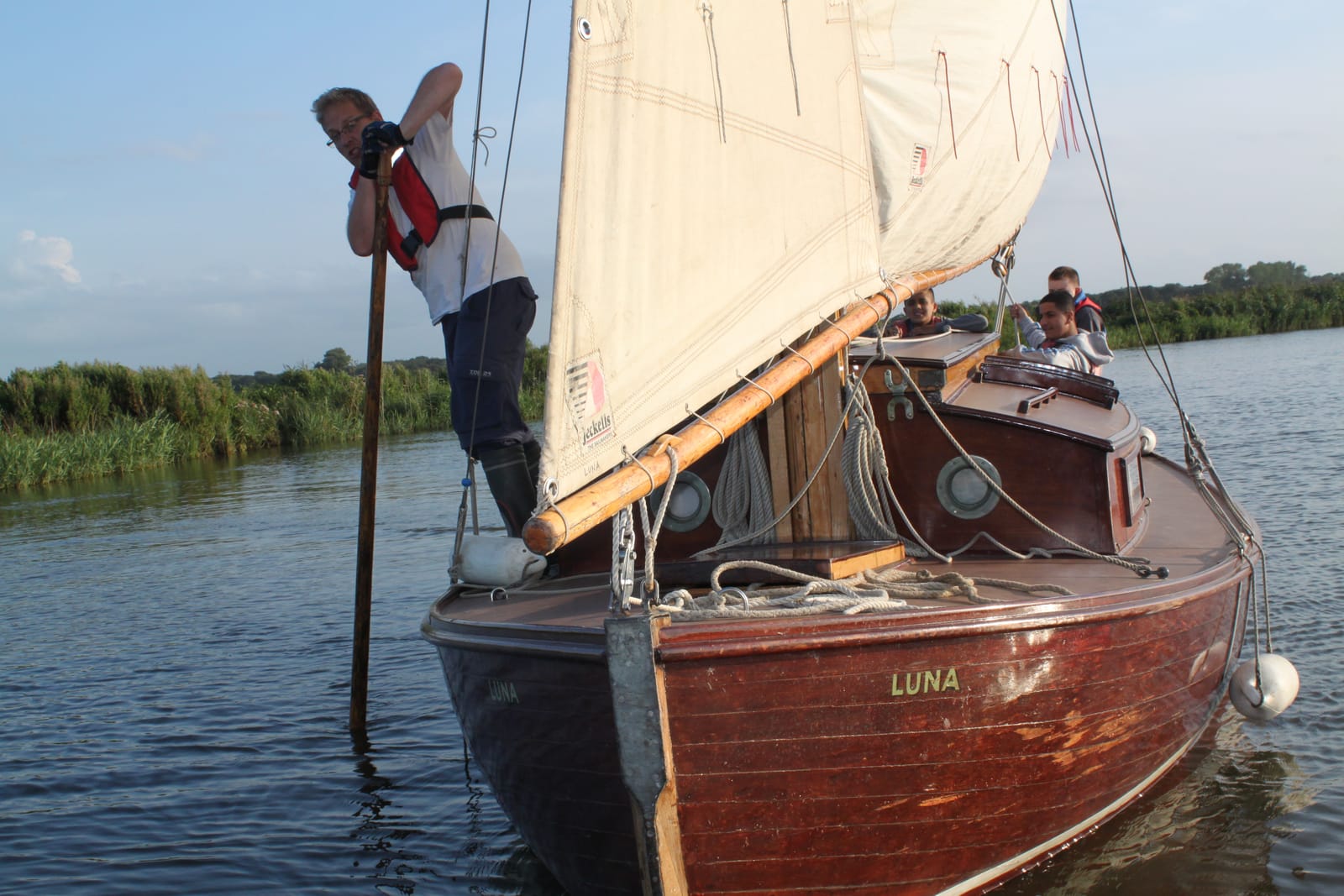 A volunteer working the bow of the yacht Luna in golden evening light, children looking out from the cabin