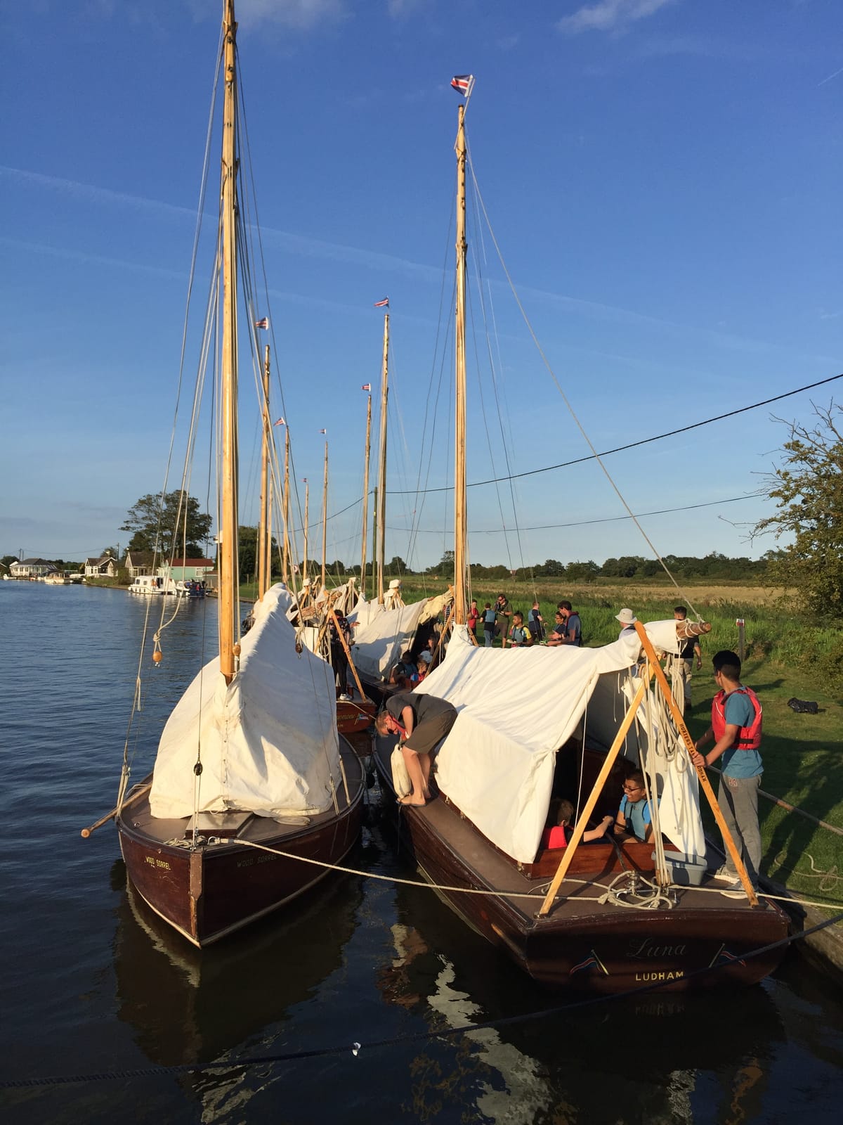 Traditional wooden half-deckers moored together at golden hour, crews and children gathered on deck and the bank