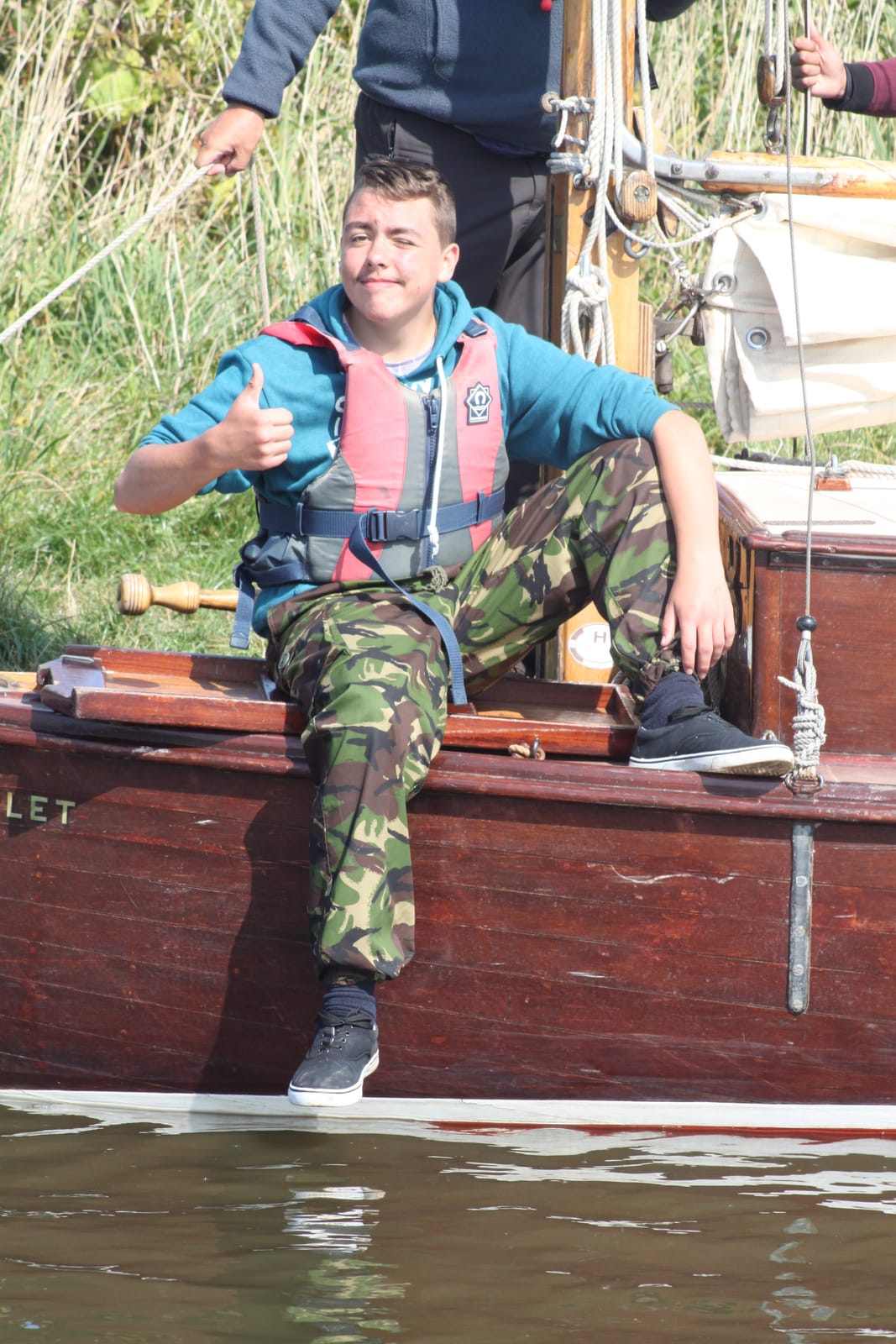 A teenage crew member sitting on the side deck of a wooden boat, giving a thumbs up
