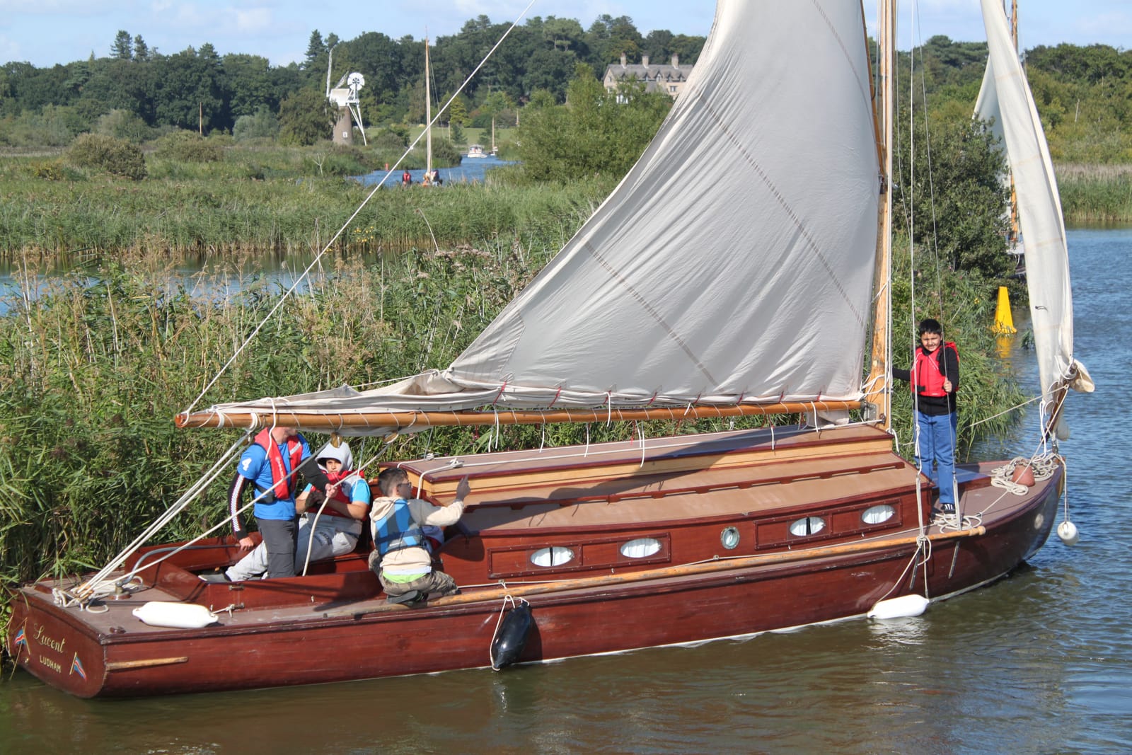 A wooden half-decker under full sail with young crew aboard, reeds and a windmill beyond